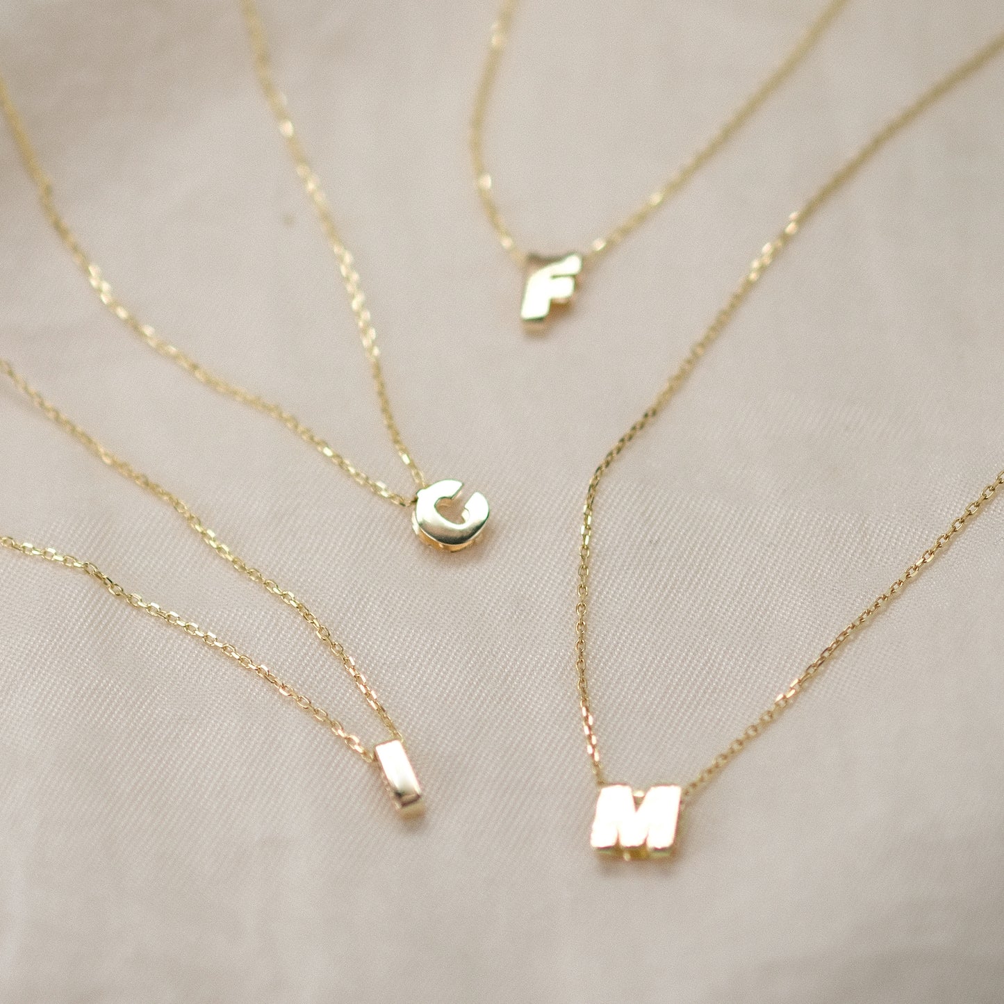 Vier Letter Chain Necklaces mit den Buchstabenanhängern I, C, F und M aus 14k Echtgold liegen auf beigem Untergrund.