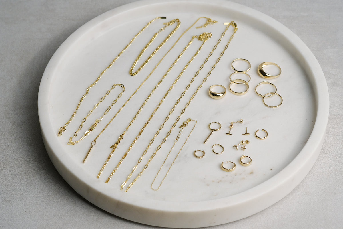 Ayu Ohrringe, Ringe, Ketten und Armbänder aus 14k massivem Gelbgold liegen auf weißem Marmor-Tablett.