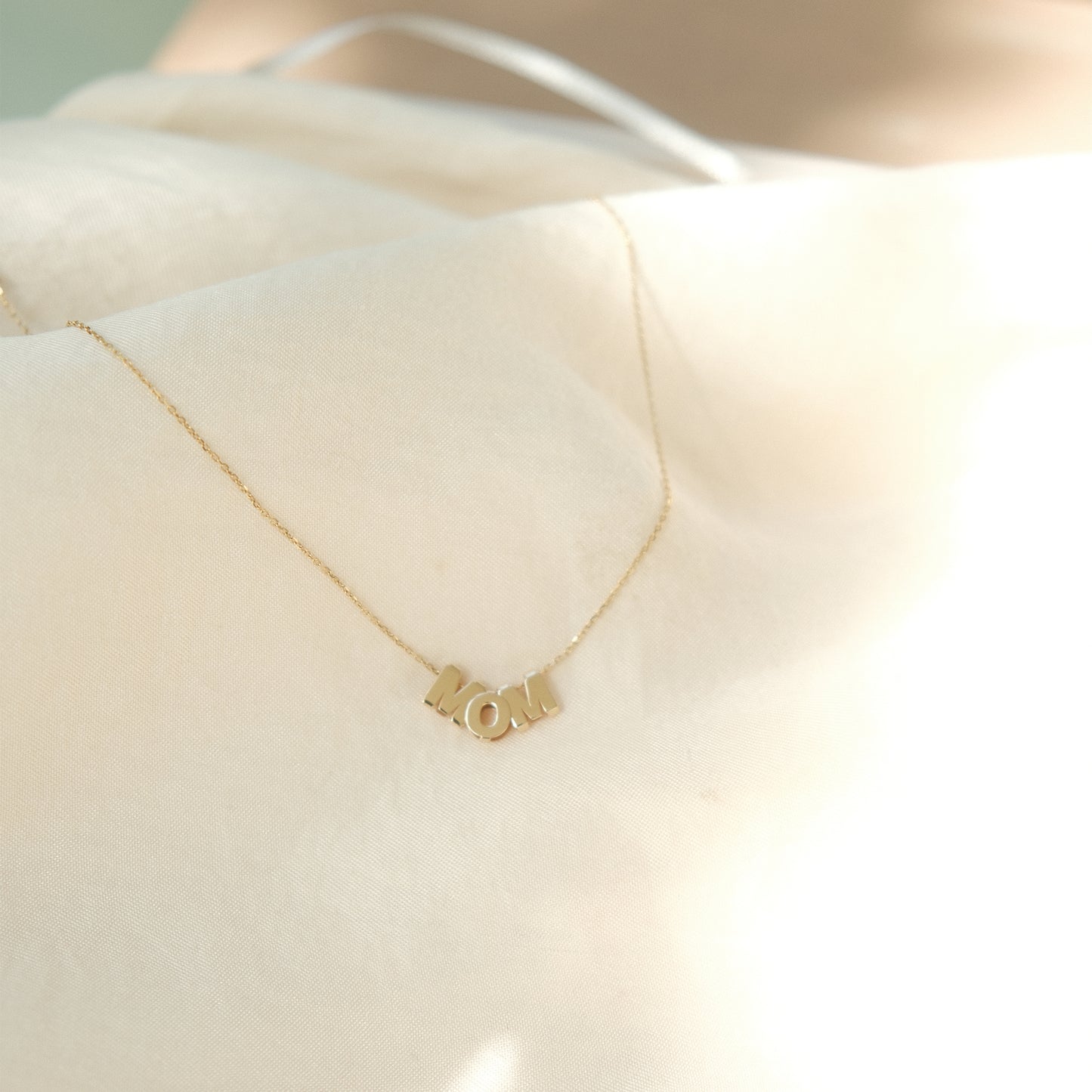 MOM-Buchstabenkette aus 14k Gold liegend auf beigem Untergrund.