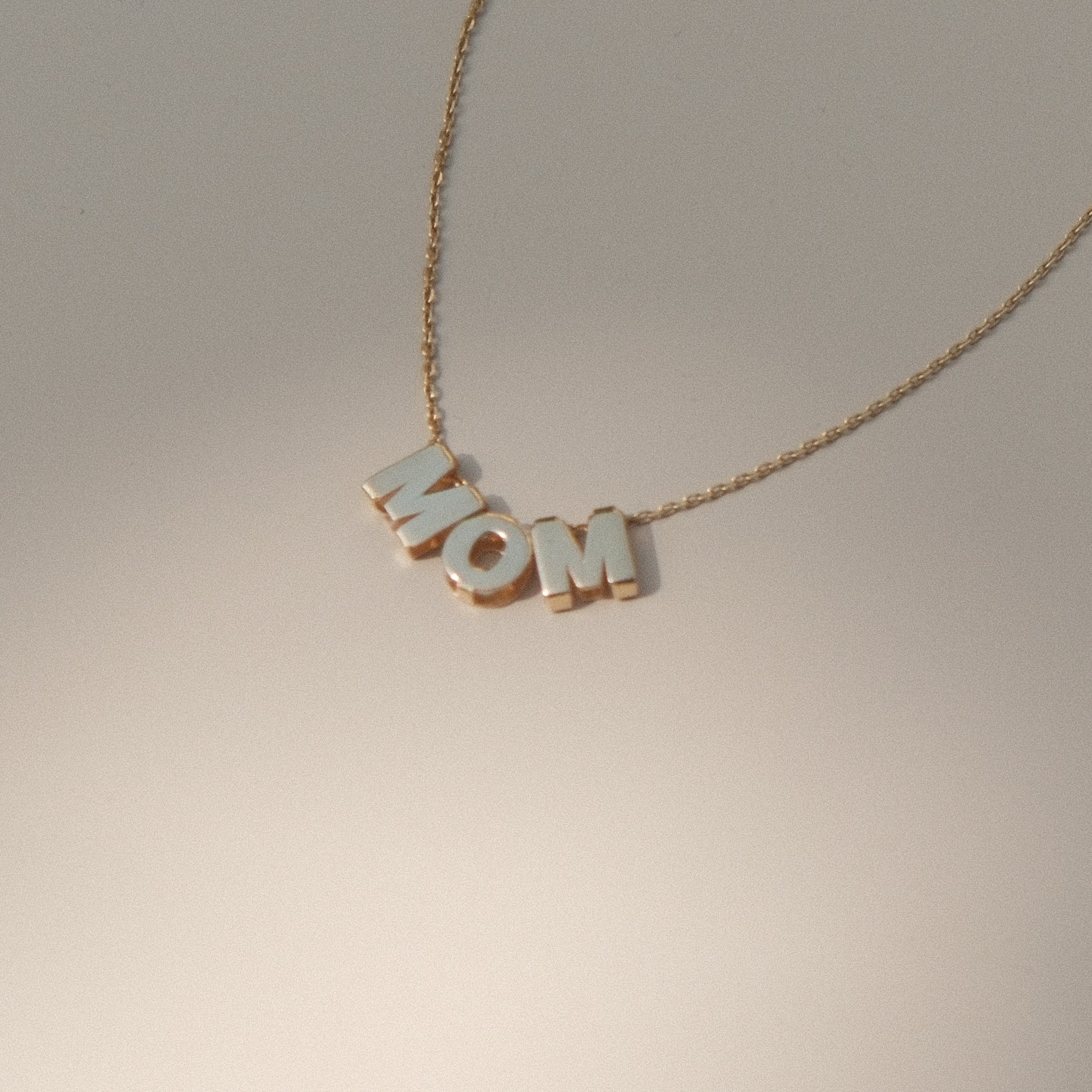 MOM-Buchstabenkette aus 14k Gold liegend auf grauem Untergrund.