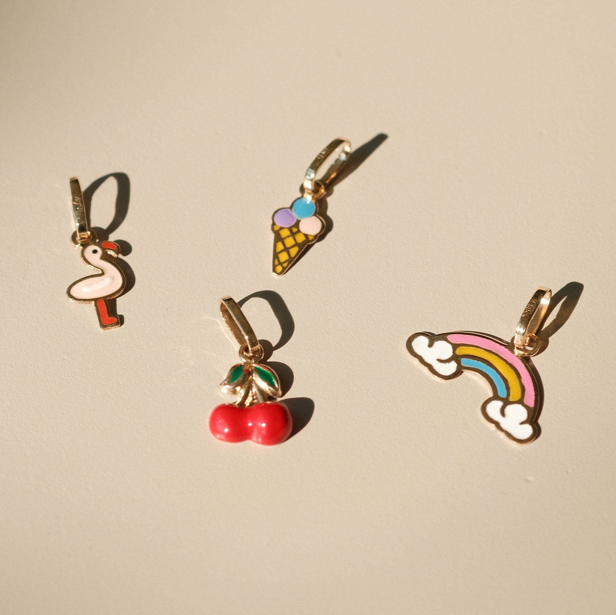 Vier bunte Charm-Anhänger aus 14k Massivgold und Emaille mit den Motiven Flamingo, Eistüte, Kirschen und Regenbogen auf beigem Hintergrund.