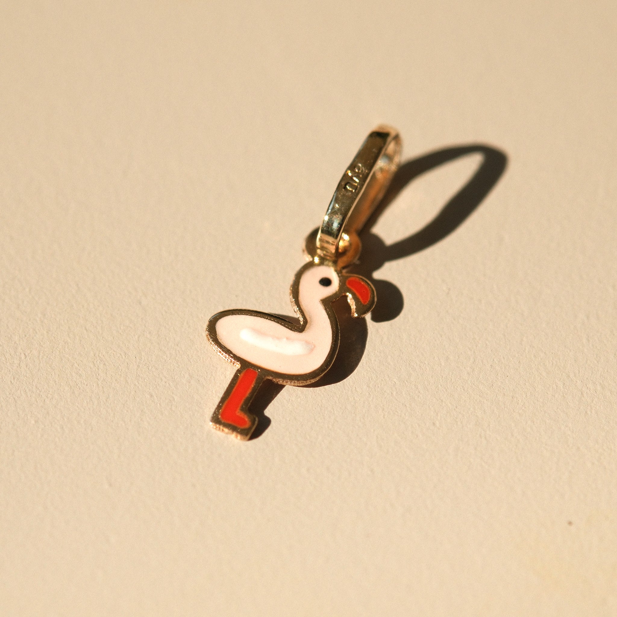 Flamingo-Anhänger aus 14k Massivgold und Emaille auf beigem Hintergrund mit Schatten.